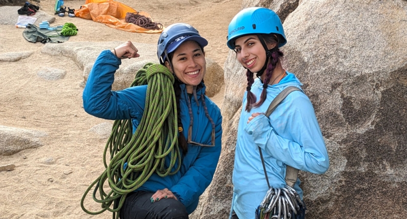 two people wearing helmets stand beside a large boulder and smile for the camera. They have climbing gear slung over their shoulders. 